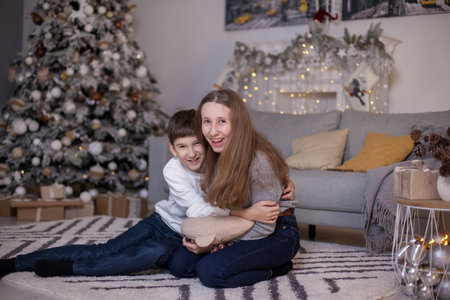 happy brother and sister sitting on floor near sofa and christmas tree with presents and looking at camera. Boy and girl at Christmas time. Merry Christmas and Happy New Yearの写真素材
