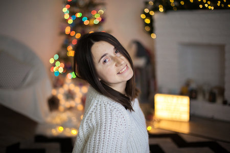 A woman in white sweater smiling and in front of christmas tree. a pretty girl with dark hair in christmas eve. Cozy holidays.の写真素材