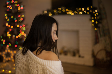 A woman in white sweater sitting backin front of christmas tree and fireplace. a pretty female with dark hair in christmas eve. Cozy holidays. Illuminated bokehの写真素材