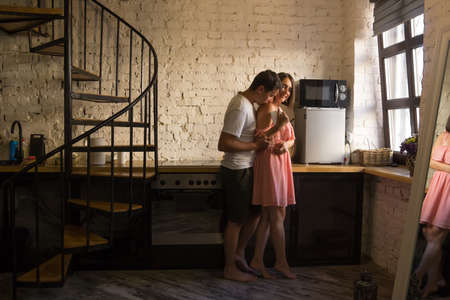 Young couple hugging on Valentines Day morning in the kitchen with spiral staircase. Barefoot people in the dinning room. Husband kisses shoulder of wife dressed in a pink dress who drinks coffeeの写真素材