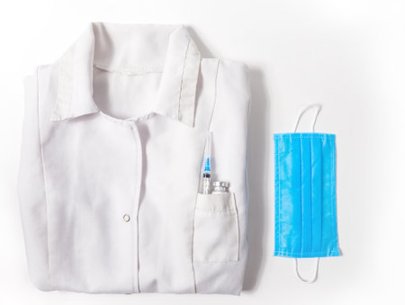 Flat lay medical gown with vaccine bottle and syringe in pocket and protective mask on white background.の写真素材