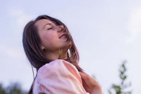 Beautiful portret of happy woman with dark hair standing in the field on the sky background in the pink dress and sensually touching her face on a warm summer evening. Joyfull summer walking.の写真素材