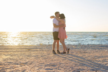 Beautiful young couple kissing on the beach at dawn. A guy in a white t-shirt hugging with a girl in a short pink dress on an empty beach near a calm sea in the early morning in summer.の写真素材