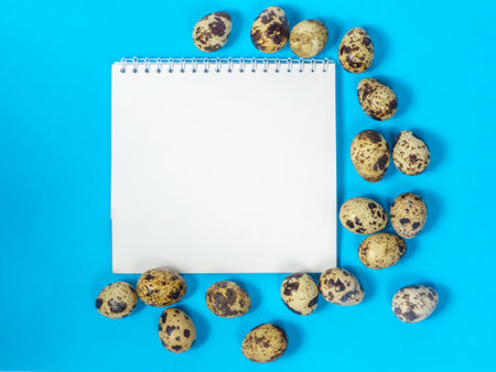 Blank sheet of paper for your text surrounded by quail eggs on blue background. Tasty, healthy food. Small raw eggs. Preparation for easter. Happy Easter Holiday card with space. Easter shopping listの写真素材