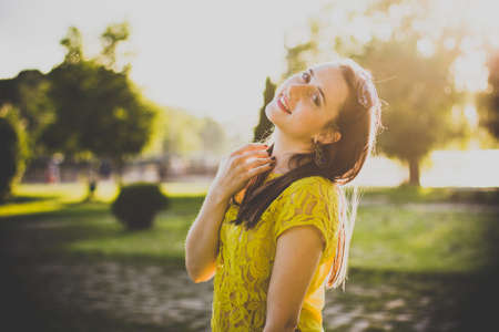 A portrait of a beautiful smiling woman with dark hair in a yellow blouse standing in the park, touching her neck with her hand. Close up photo of a girl with back sunlight on the natureの写真素材