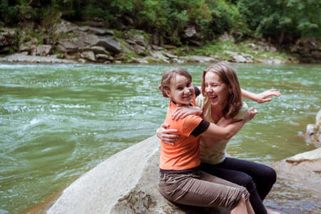 Beautiful sisters have fun while sitting on the rock near mountains river. Two girl hugging and smiling on the big stone in forest in front of the big green river. Happy summer family vacation.の写真素材