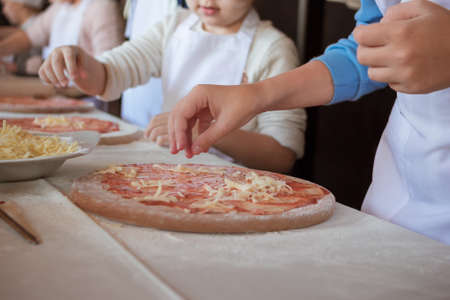 Cooking class, culinary. Kids hand puts cheese on a row pizza on a round wooden cutting board sprinkled with flour on the white table. Children in uniform preparing food, meal in the kitchenの写真素材