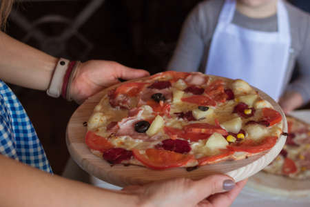 Cooking class, culinary. Womans hands holding a baked pizza with tomatos, cheese, bacon, salami, corn and olives on a round wooden cutting board. Children preparing food, meal in the kitchenの写真素材