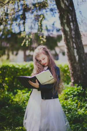 Little beautiful girl in dress and black jacket holding a book in spring garden. Portrait of child...