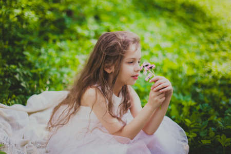A girl in white dress sniffs a bouquet of daisies in spring cherry garden. Portrait of child among white flowers trees. Young lady sitting in sunny blooming park with green grass and yellow dandelionsの写真素材