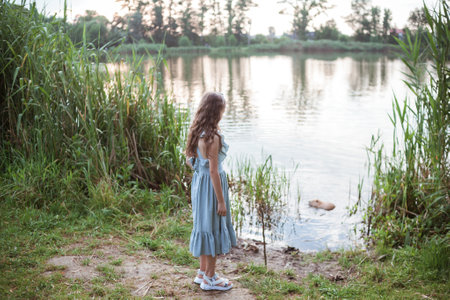 A girl in a long blue dress stands on the shore of the lake and looks at nutria in the water. Child watching with interest on coypu swimming. Young lady and Myocastor coypus in public park in summer.の写真素材