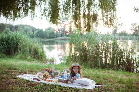A girl in a long summer dress with long hair is on a white blanket with fruits and pastries, white basket with flowers. Concept of having picnic in a city park during summer holidays or weekends.の写真素材