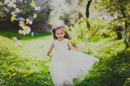 Little girl with long hair in beautiful white dress running in spring cherry garden. Happy child among white flowers trees. Childhood. Young lady playing in sunny blooming park at springtime.の写真素材