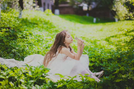 A girl in white dress sniffs a bouquet of daisies in spring cherry garden. Portrait of child among white flowers trees. Young lady sitting in sunny blooming park with green grass and yellow dandelionsの写真素材