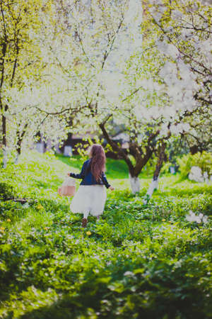 Little girl in black jacket and white dress running away with pink backpack in spring cherry garden. Happy child among white flowers trees. Young lady walking in sunny blooming park at springtimeの写真素材
