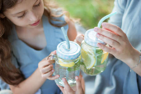 Cropped photo of a woman and a girl in blue dresses sitting and drinking lemonade in glass mugs during rest near the river. Concept of having family picnic in a city park on summertime.の写真素材