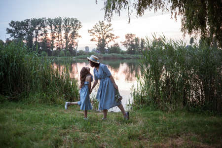 Woman posing outdoor in the nature with bouquet of flowers and the gorgeous view of lake and sun. the mother having fun with girl or daughter. Models dressed in blue dresses. Happy mothers dayの写真素材
