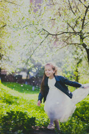Little girl in black jacket and white dress dancing in spring cherry garden. Happy child among white flowers trees. Childhood. Young lady enjoys walking in sunny blooming park at springtime.の写真素材
