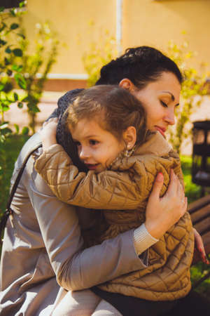 A little girl with dark hair in a mustard-colored jacket hugs her mother sitting on bench in a park. Mom embracing her lovely daughter on the warm autumn day. Motherhood. Happy mothers dayの写真素材