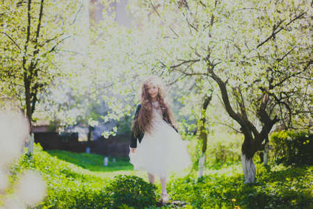 Little girl in black jacket and long white dress standing in spring cherry garden. Happy child among white flowers trees. Childhood. Young lady walking in sunny blooming park at springtime. Soft focusの写真素材