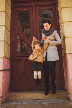 Little happy girl playing with her mom near old brown wooden door. Smiling woman in gray trench and scarf jumping with her daughter from the stairs. Concept of family relationship and motherhood.の写真素材