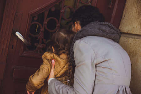 Little girl with her mom looking out the window of old brown wooden door. Woman in gray trench and scarf holding her daughter on the stairs. Concept of family relationship, motherhood and curiosity.の写真素材