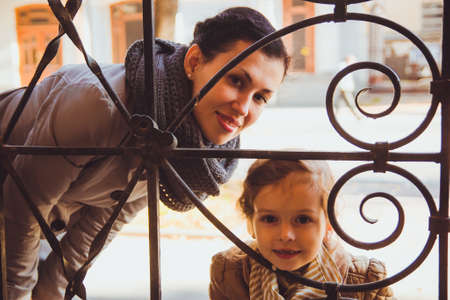 Little girl with her mom looking out the window of old ornate iron door. Woman in gray trench...