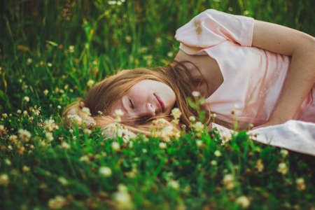 Portrait of a girl with long hair in a pink dress lies and sleeps in the green grass with white...
