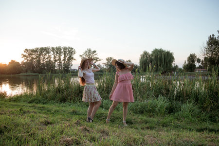 Two young beautiful smiling women in short summer dresses and straw hats posing in front of lake on warm summer day at sunset. Best girlfriends outdoors. Positive models having fun in the parkの写真素材