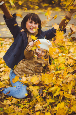 Beautiful young mother and her happy daughter are having fun in the forest at autumn. Walks in an autumn park with the kids. Family spending great time playing together with fallen leaves.の写真素材
