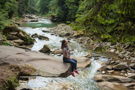 Happy smiling girl in a white t-shirt, blue jeans and red sneakers sitting near mountain river. Young woman enjoys fresh air in forest resting on a rock above a waterfall. lonely travel, side viewの写真素材