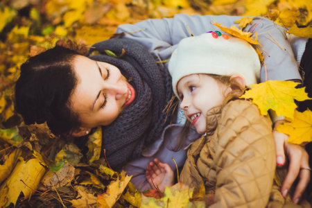Beautiful young mother and her happy daughter lying on the ground in the forest at autumn. Walks in an autumn park with the kids. Family spending great time playing together with fallen leaves.の写真素材
