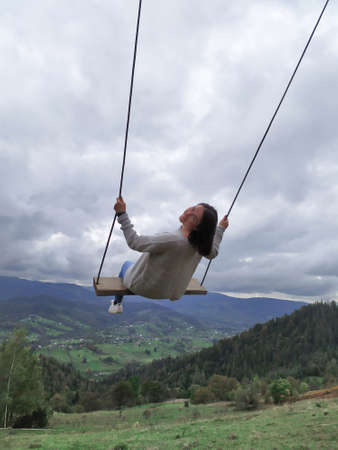 Back of girl with dark hair in a gray sweater sitting on a swing with the mountain view. Calm and...