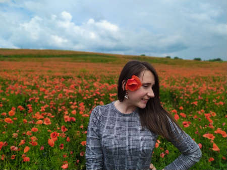 Smiling woman in a long gray dress standing in the middle of the red wildflowers field and cloudy sky background. Happy brunette girl in a red field of poppies stands and enjoys life. Beauty in natureの写真素材
