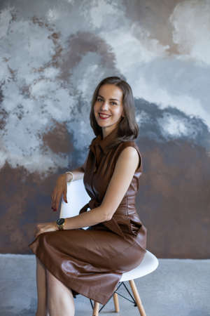 Portrait of a beautiful young business woman with brown hair in a brown dress. Side view of happy smiling girl with red lips on gray background with copy space. The concept of business and success.の写真素材