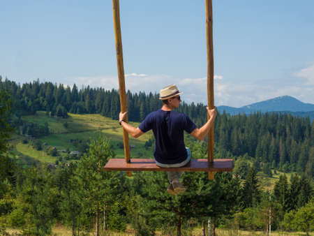 Back view of young man sitting on a swing on a background of beautiful nature with the mountain range. Calm and quiet wanderlust concept moment when person feels happiness, freedom and life.の写真素材