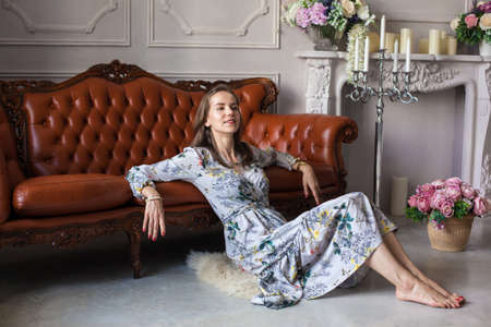 Young beautiful woman in floral dress sits on floor near brown leather sofa surrounded different flowers and candles on white wall background. Happy smiling girl rests at home and looking at camera.の写真素材