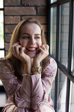 Beautiful young woman in pink dress resting on the windowsill. Portrait of happy smiling girl sits...