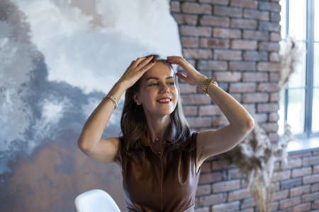 Portrait of a beautiful young business woman with brown hair in a brown dress. Happy smiling girl with red lips sits in white chair on gray and brick background. The concept of business and success.の写真素材