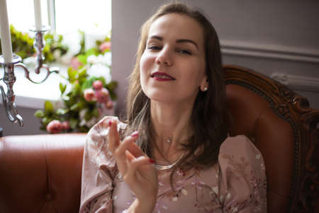 Young beautiful woman in pink dress sits on brown leather sofa with candelabrum on her side on white wall background. Happy smiling girl resting at home. Lady is on romantic date in beautiful interiorの写真素材