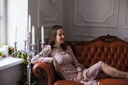 Young beautiful woman in pink dress sits on brown leather sofa surrounded different flowers and candles on white wall background. Happy smiling girl resting at home and looking at window. Spring mood.の写真素材