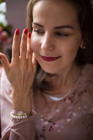 Young beautiful woman in pink dress and stylish accessories sits on brown leather sofa on white wall background. Portrait of happy smiling girl with red lips resting at home and looking at camera.の写真素材