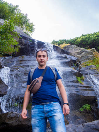 Tourist rises in the mountains to the highest waterfall of Ukraine under the mountain Goverla. Man stands on the background with waterfall of wild mountain slopes of the Carpathian Mountains.の写真素材