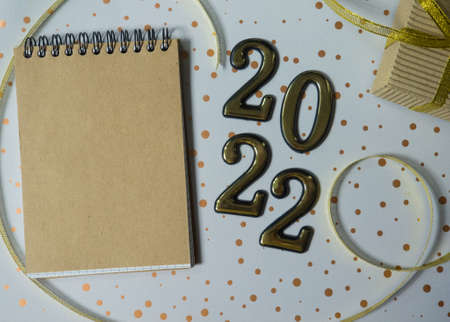 2022 and notebook with empty place for text on the white dotted background. gold figures lie next to a small gift in craft brown wrapper and with long shiny yellow ribbon. Christmas and New year cardの写真素材
