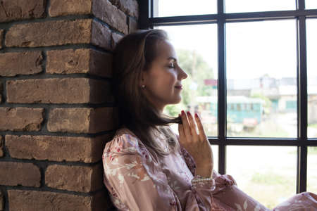 Beautiful young woman in pink dress resting on the windowsill. Portrait of happy smiling girl sits...