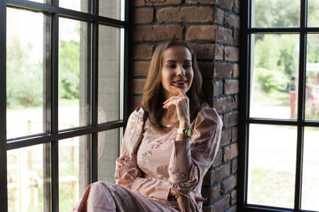 Beautiful young woman in pink dress resting on the windowsill. Happy smiling girl sits on the...