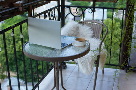 Laptop and cup of coffee on the table on the cozy balcony with flowers at home with city view. Remote work, doing business on vacation. Fast Internet, communication on weekends. Always in touch.の写真素材