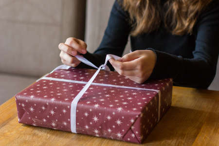 Close up view of a girl in black clothes ties a bow with white ribbon on a Christmas gift. Teen girl packing a christmas present on wooden table at home. Concept of shoping for holidays.の写真素材