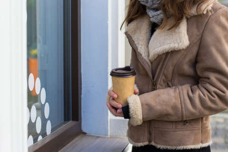 A young girl with blond hair in a brown coat stands near showcase on the street with takeaway coffee in cold winter day. Close up photo with hands holding paper cup with coffee. Copy spaceの写真素材