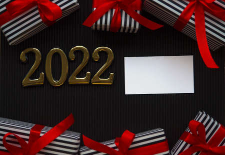 2022 on black background. 2022 gold figures lie near gifts in a stripped black and white wrapper and with red ribbons on the dark corrugated cardboard. Christmas and New year card with place for textの写真素材